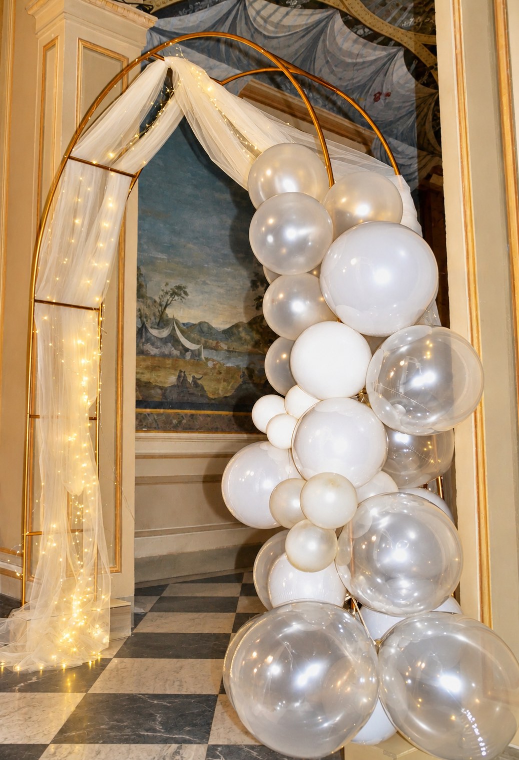 Arco dorato con tulle, luci e palloncini bianchi in una sala affrescata